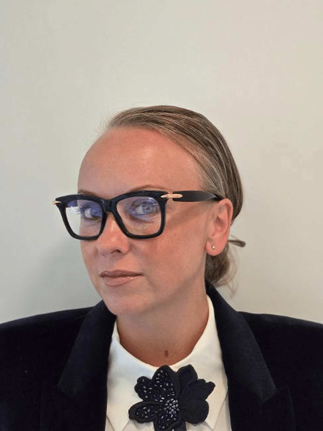 Woman with sleek hair and statement glasses wearing a formal outfit, featuring a white collar and black flower brooch,